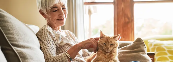 Une femme souriante caresse un chat roux sur ses genoux, sur le canapé.