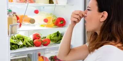 Femme qui se bouche le nez devant un frigo ouvert rempli d'aliments variés.