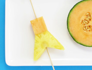 Une brochette en bois avec des morceaux d'ananas et de melon, et un melon coupé en deux sur un plateau blanc.