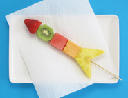Une brochette aux fruits qui ressemble à une fusée sur un plateau blanc et une feuille d'essuie-tout Okay.