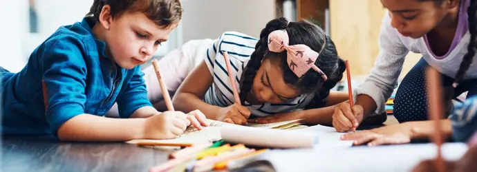 Trois enfants allongés sur le ventre par terre, faisant du coloriage avec des crayons de couleur.