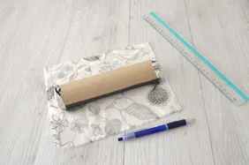 Tube de rouleau d’essuie-tout avec une fermeture éclair, posé sur un carré de tissu, à côté d’un stylo bleu et d’une règle.