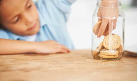 Een kind reikt in een glazen koekjespot op een houten tafel om een koekje te pakken
