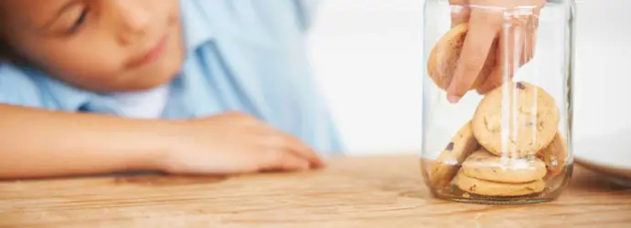 Een kind reikt in een glazen koekjespot op een houten tafel om een koekje te pakken