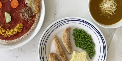Pizza décorée avec des légumes, un nez  concombre, des yeux carottes, un sourire maïs, et à coté une assiette de petits pois et des nuggets et un bol de soupe.