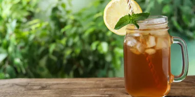 Mug en verre transparent posé sur une table en bois dans un jardin, dans lequel se trouve du thé glacé à la menthe. Il y a une tranche de citron sur le bord du mug, et des plantes en arrière-plan. 