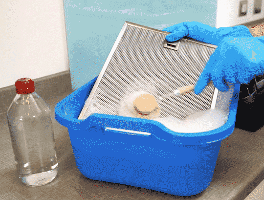 Une personne frotte un filtre avec une brosse dans un bassin plein d'eau chaude savonneuse et de vinaigre blanc.
