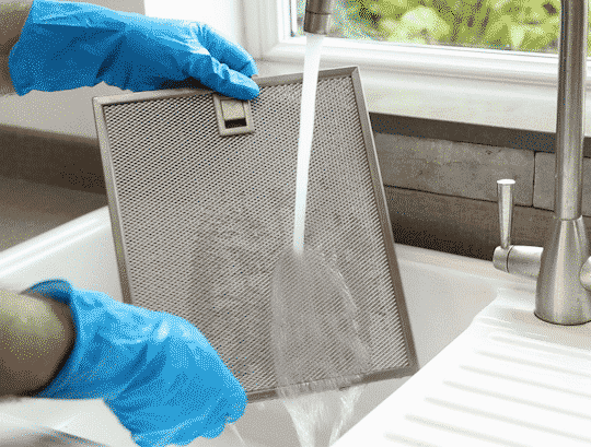 Une personne qui porte des gants bleus lave un filtre sous un jet d'eau dans un lavabo blanc.