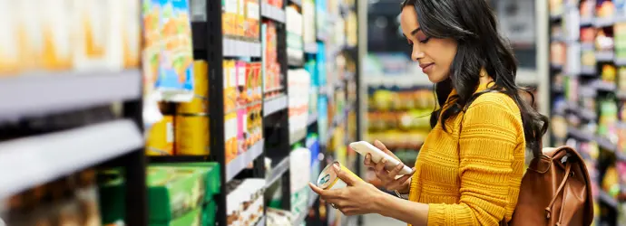 Une femme tient une boîte de conserve dans une main et son téléphone dans l'autre, dans un supermarché.