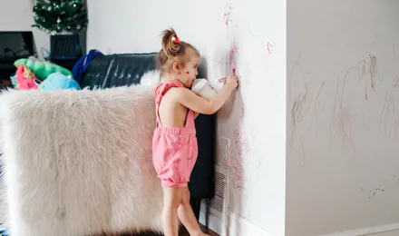 Petite fille habillée en rose dans un salon. Elle dessine au feutre sur le mur d'une main, et boit du lait de son biberon de l'autre main.  