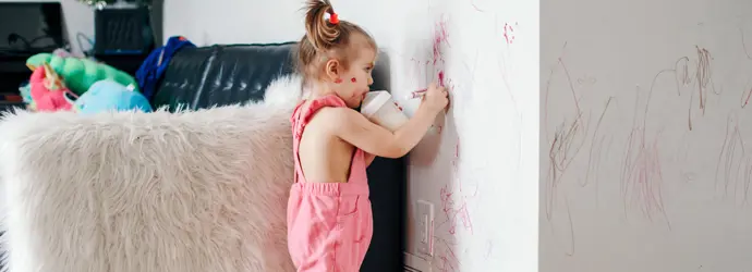 Petite fille habillée en rose dans un salon. Elle dessine au feutre sur le mur d'une main, et boit du lait de son biberon de l'autre main.
