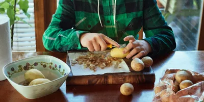 Jeune garçon épluchant des pommes de terre sur une table en bois dans une cuisine.