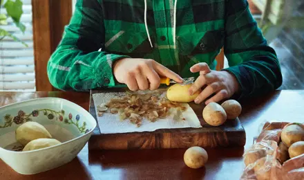 Jeune garçon épluchant des pommes de terre sur une table en bois dans une cuisine.