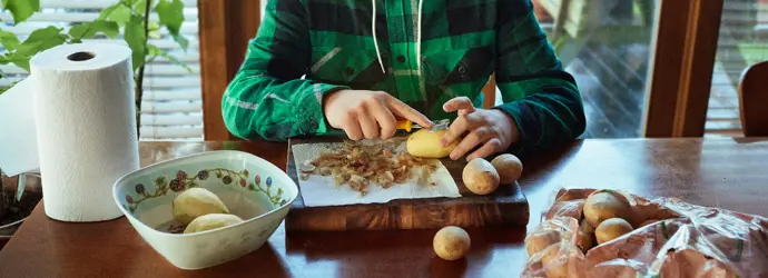 Jeune garçon épluchant des pommes de terre sur une table en bois dans une cuisine.