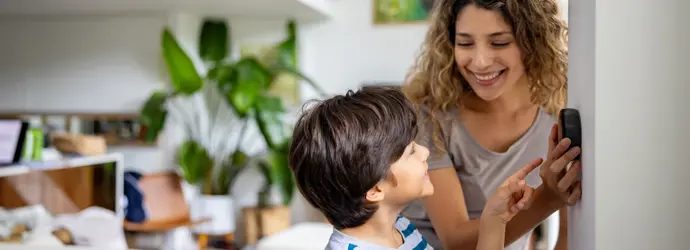 Une mère aux cheveux bouclés et un jeune garçon souriant changent un thermostat.