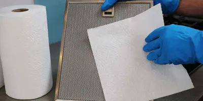 Une personne qui porte des gants bleus en caoutchouc tient le filtre d'une hotte et un papier essuie-tout.