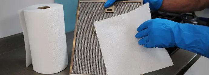 Une personne qui porte des gants bleus en caoutchouc tient le filtre d'une hotte et un papier essuie-tout.